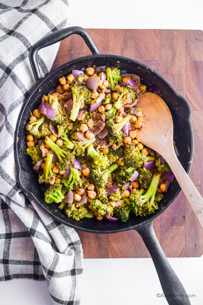 broccoli chickpea stir fry in a skillet with wooden spoon and kitchen towel beside pan