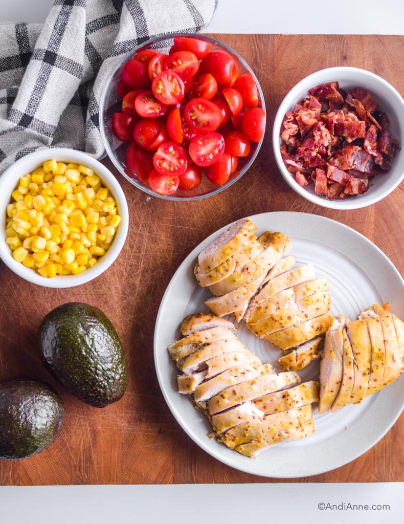 Plate of sliced chicken, two avocados, bowl of corn, bowl of sliced cherry tomatoes and bowl of chopped bacon.