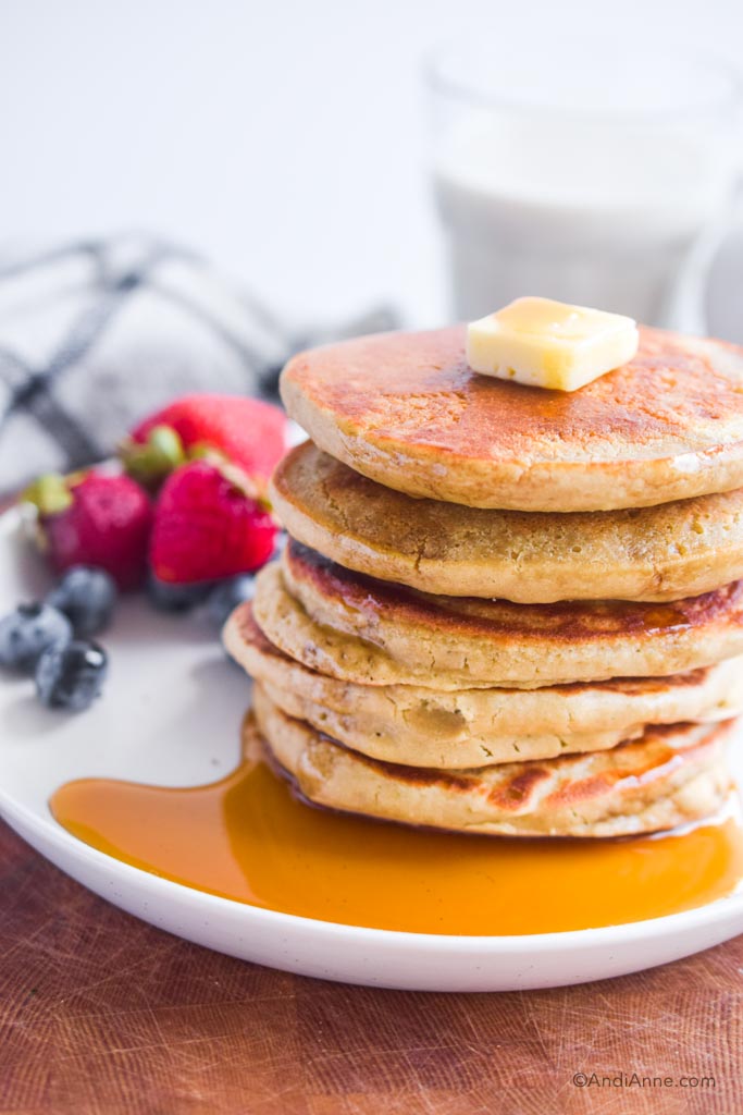stack of quinoa flour pancakes with butter and maple syrup poured on. Berries in background