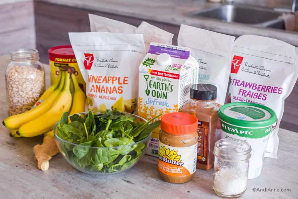 ingredients to make smoothie packs on a counter, including frozen fruit bags, a bowl of spinach, bananas, milk, cinnamon, yogurt and nut butter