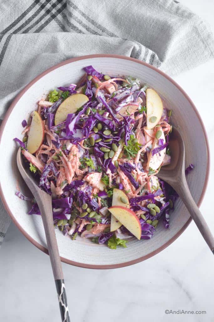 Carrot apple cabbage coleslaw in a white bowl with wood serving spoons. Kitchen towel beside it.