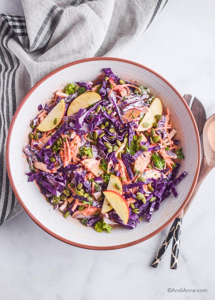 Carrot apple cabbage coleslaw in a white bowl with serving spoons on the side. 