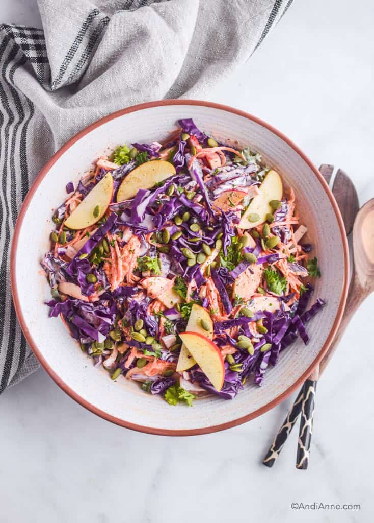 Carrot apple cabbage coleslaw in a white bowl with serving spoons on the side.