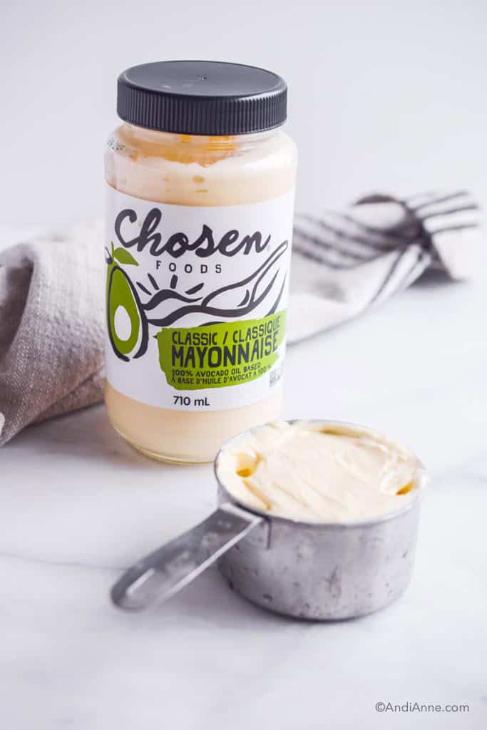jar of chosen foods avocado mayonnaise with measuring cup in front and kitchen towel in back
