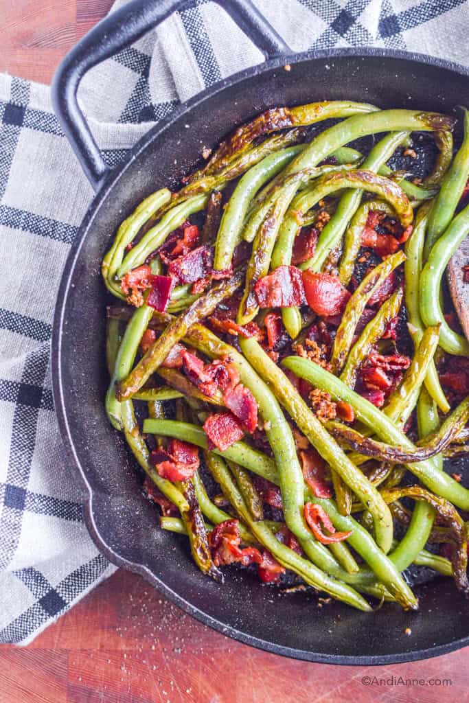 Cooked green beans and bacon in black skillet with plaid kitchen towel underneath
