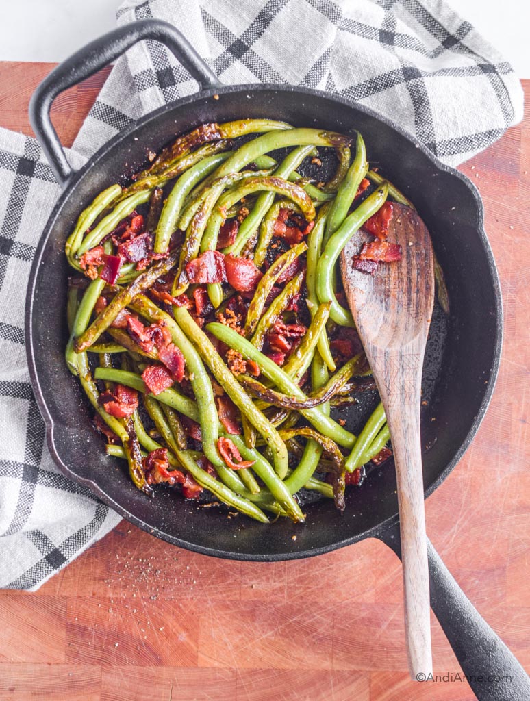 garlic green beans with bacon in a black skillet with wooden spoon and kitchen towel behind it
