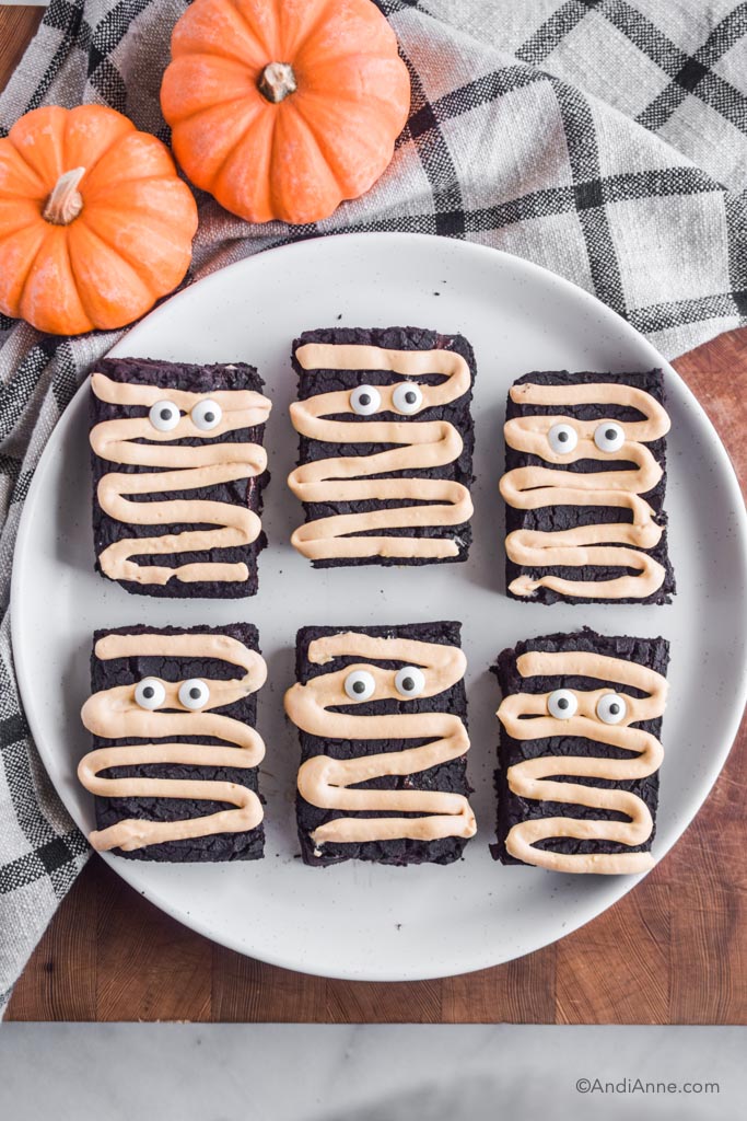 Halloween mummy chickpea brownies on a big white plate on wood block. Plaid kitchen towel and small pumpkins surround the plate.