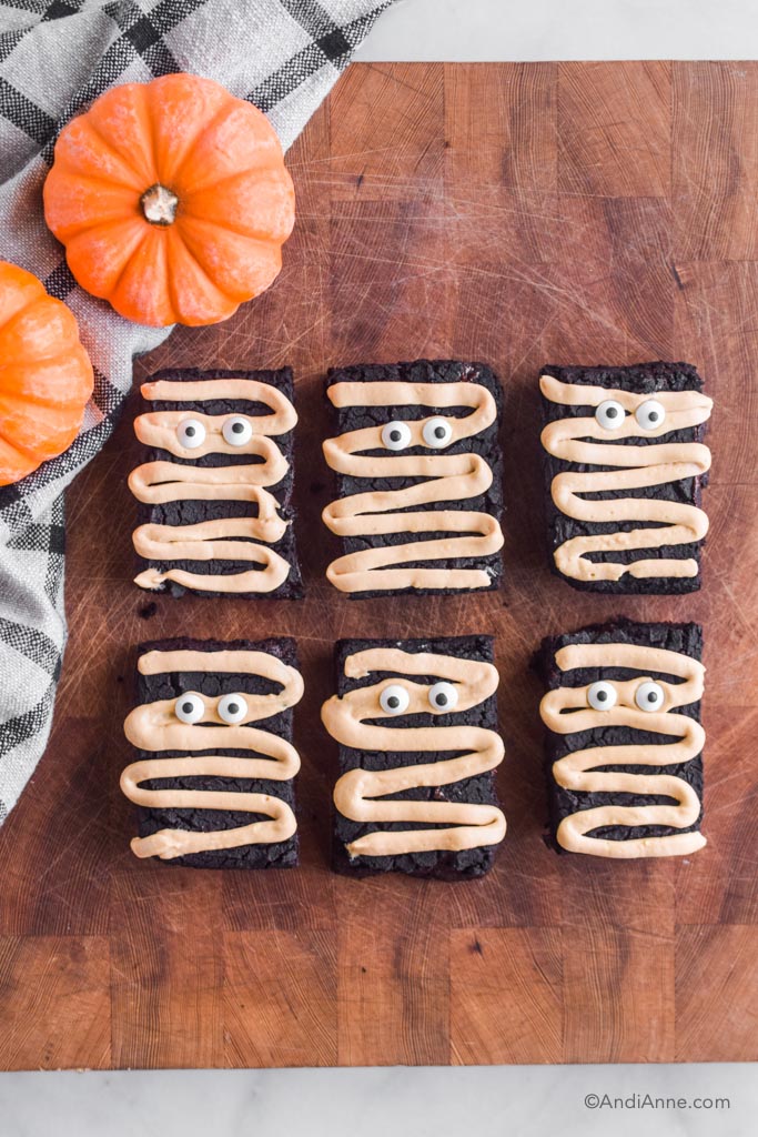Six brownies with mummy designs on a wood butcher block with small pumpkins and kitchen towel on the left side.
