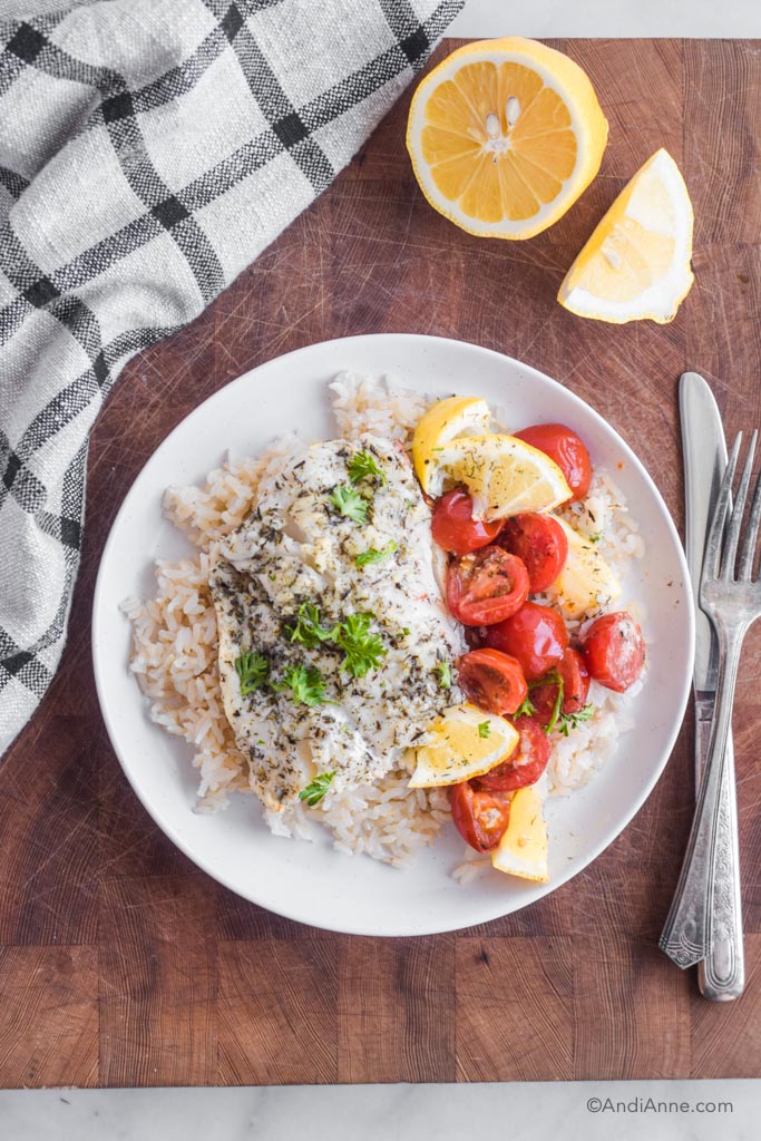 Baked cod, lemon wedges, and cherry tomatoes on top of a bed of rice on a white plate. Fork and knife beside it. 
