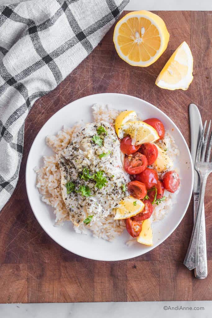 Baked cod, lemon wedges, and cherry tomatoes on top of a bed of rice on a white plate. Fork and knife beside it.