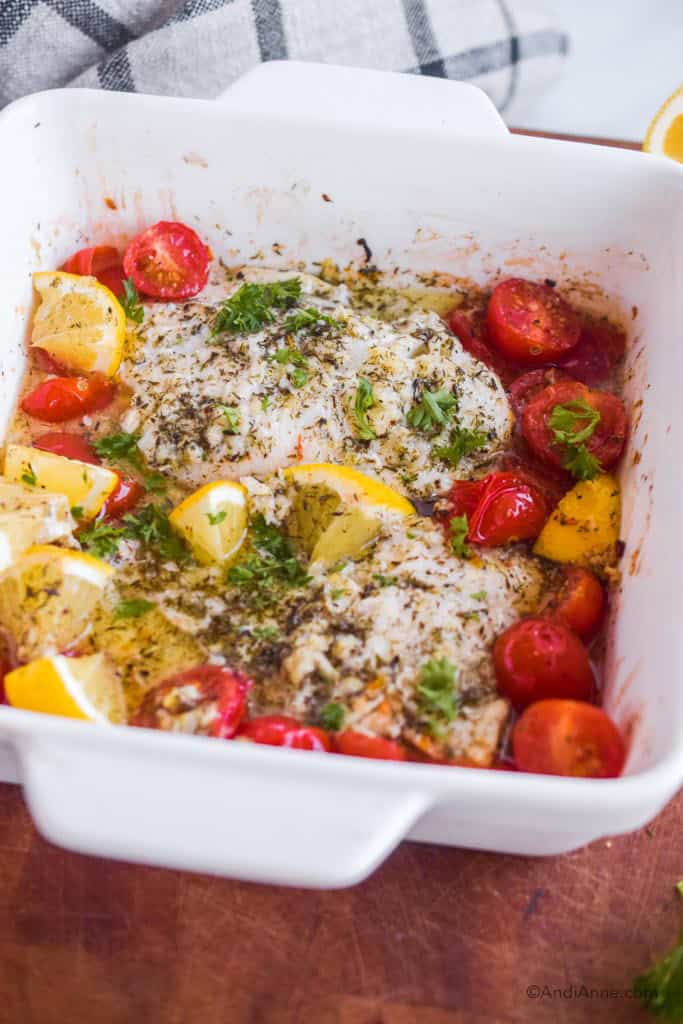 Side angle close up of white dish with baked cod, cherry tomatoes, and lemon slices. All coated in an herb dressing.