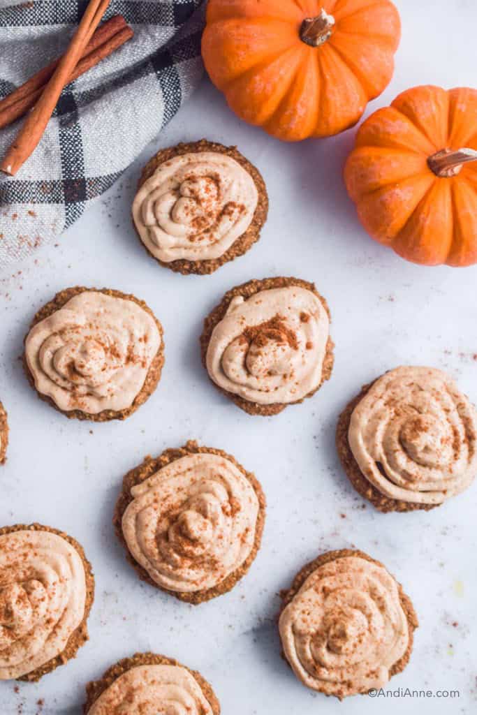pumpkin oatmeal cookies wiht cream cheese frosting and sprinkled cinnamon on a white counter. Two small pumpkins, cinnamon sticks and a kitchen towel beside them.