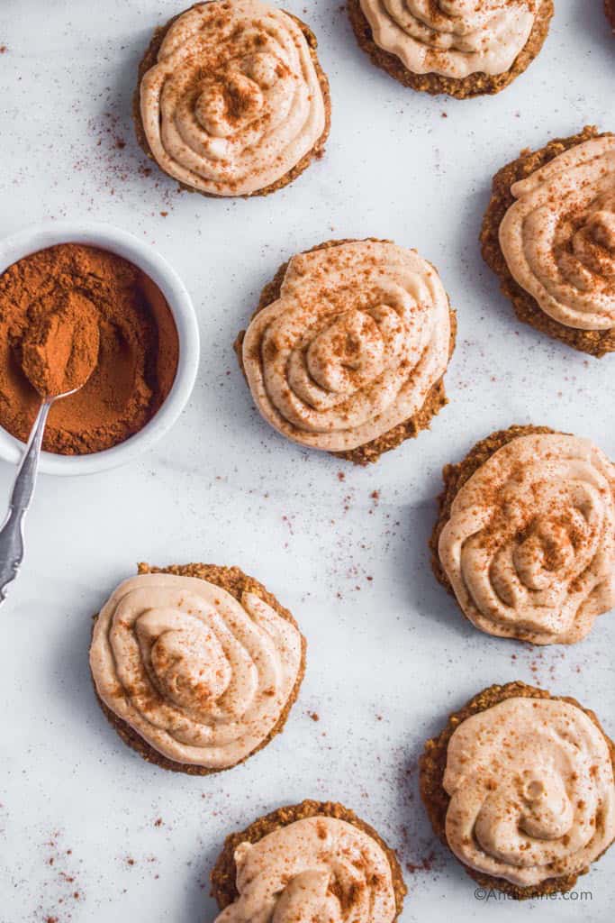 cookies with cream cheese frosting and a bowl of cinnamon with small spoon on white marble counter