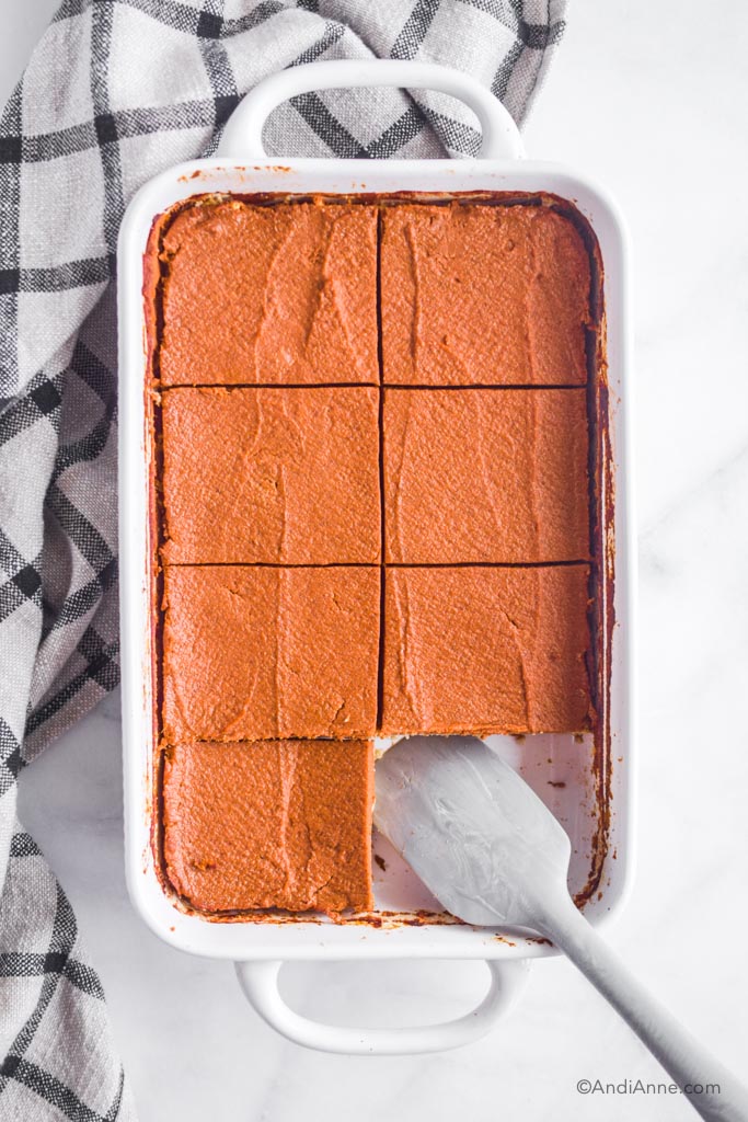 cooked and sliced pumpkin pie bars sitting in white baking dish. Grey spatula at the bottom with one slice removed.