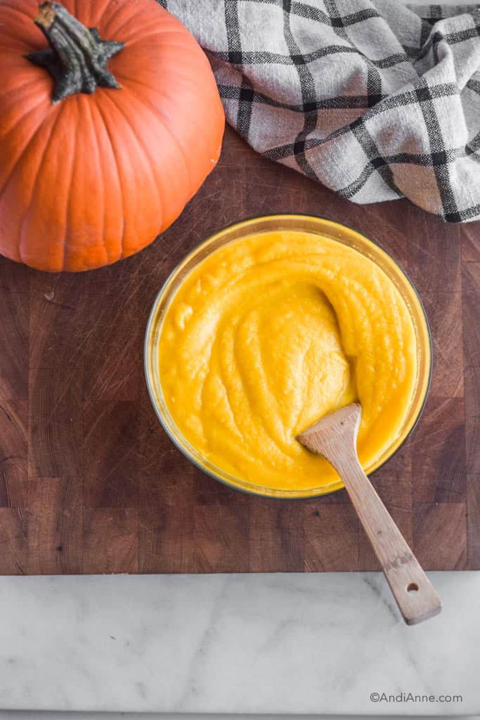 homemade pureed pumpkin in a bowl with wood spoon. Bowl on wood cutting board with kitchen towel and small pumpkin beside it.