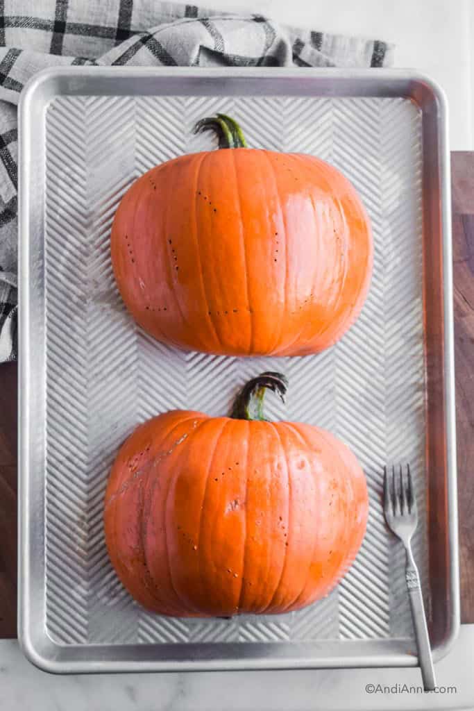 pumpkin cut in half and poked with a fork lying face down on a baking sheet. Fork beside it.
