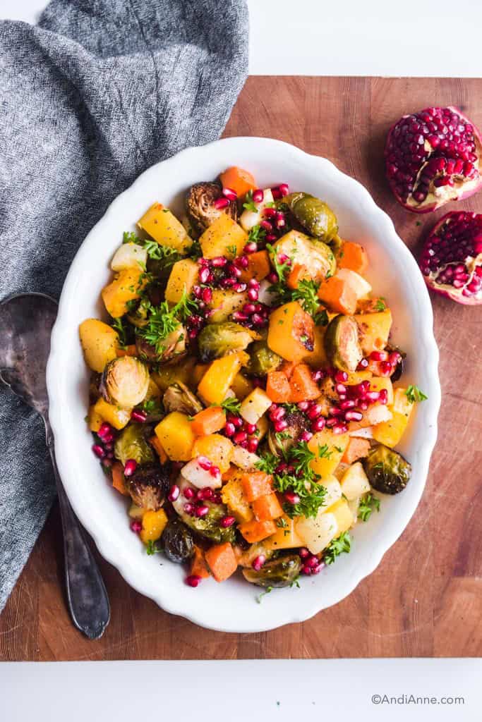 roasted root vegetables with pomegranate seeds in white serving dish on wood cutting board