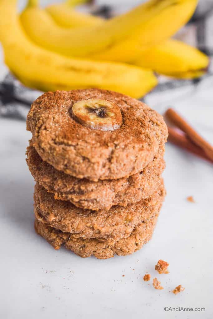 Four stacked banana bread cookies. Bananas and cinnamon in background.