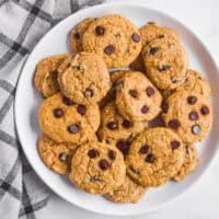Pile of cookies stacked on a white plate with kitchen towel on left side.