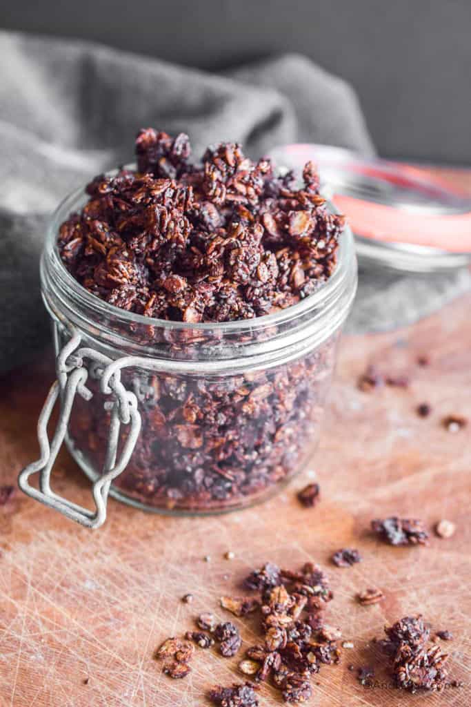 chocolate granola in a glass jar with small clumps spilled on the wood counter in front.