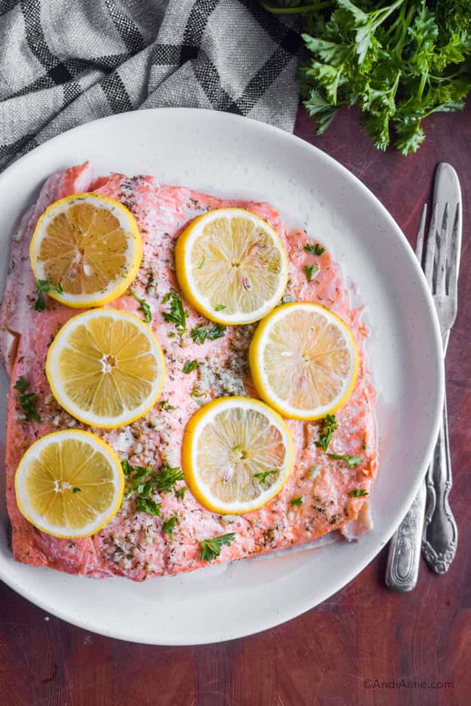 Looking down at a plate with steelhead trout filet and slices of lemon on top. Fork and knife and parsley beside the plate.