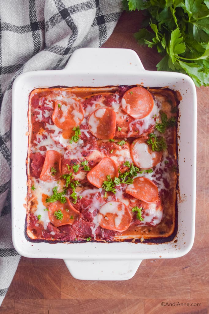 low carb pizza chicken in a white square baking dish with kitchen towel and parsley surrounding it.