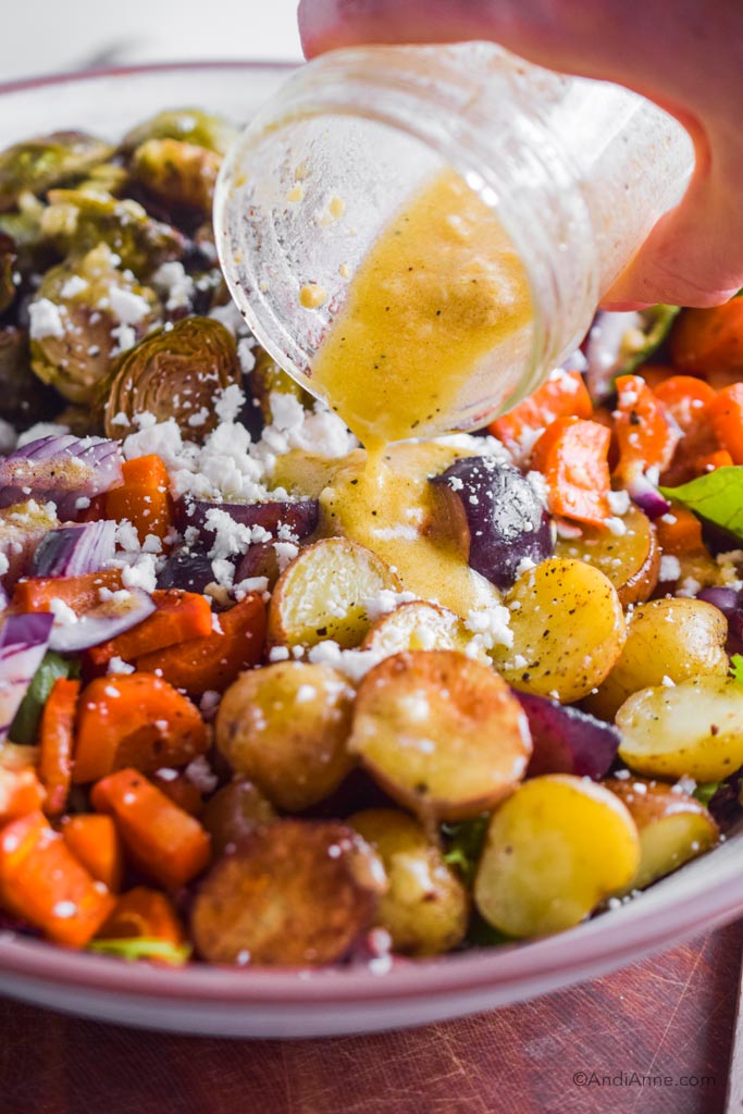 A hand pouring salad dressing overtop of the roasted vegetables in the salad bowl.