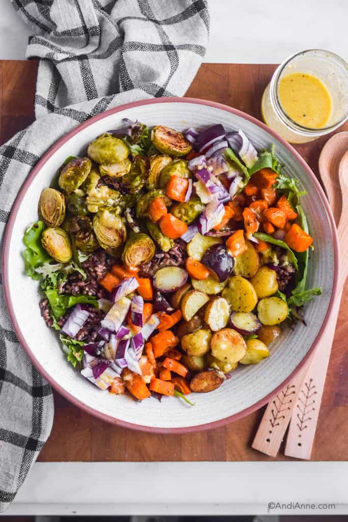 Roasted vegetable wild rice salad in a white bowl. Salad dressing in mason jar and wooden spoons on the side of the bowl.