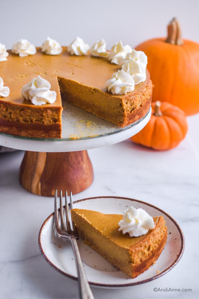 sugar-free pumpkin cheesecake on a cake stand with pumpkins in the background. Slice of cheesecake on a small white plate in the front with a fork.