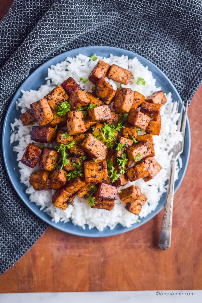 basil balsamic pan fried tofu on a blue plate with white rice and chopped parsley. A black kitchen towel and fork are beside the plate.