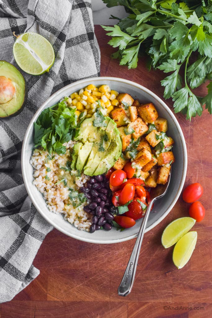 crispy tofu burrito bowl with a fork surrounded by parsley, lime and tomatoes.