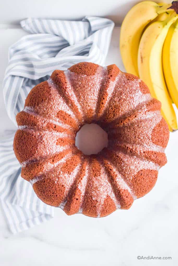 looking down at banana bundt cake with sugar glaze on top. Bananas and kitchen towel beside it.