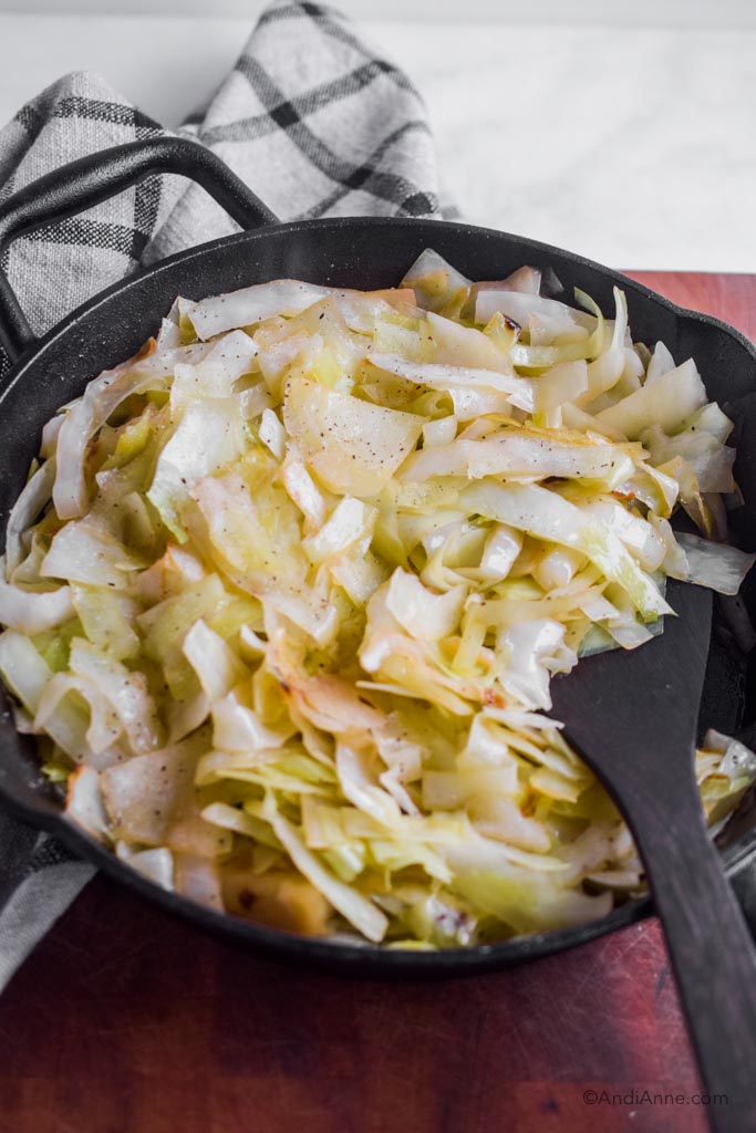 fried cabbage in a black skillet with wooden spoon. Kitchen towel behind the pan.