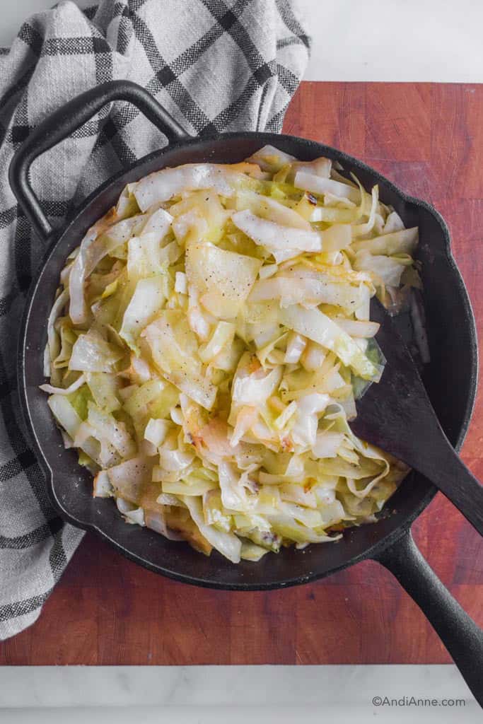 Looking down at a black skillet with fried cabbage and wooden spatula. Kitchen towel beside the pan. Pan is on a wood butcher block.