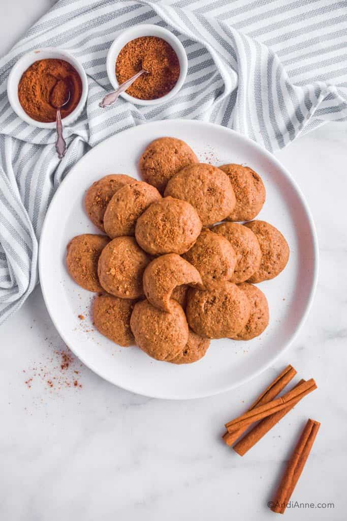 plate of applesauce spice cookies with small bowls of spices, striped kitchen towel and cinnamon sticks surrounding it.