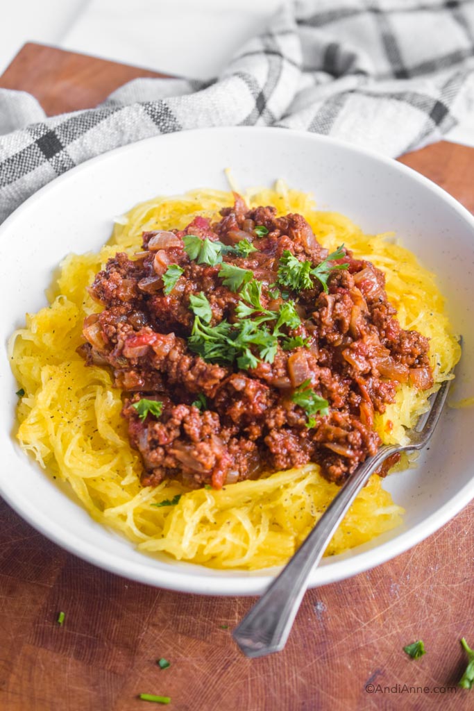 spaghetti squash and bolognese sauce in a white bowl with chopped parsley and a fork on the side.