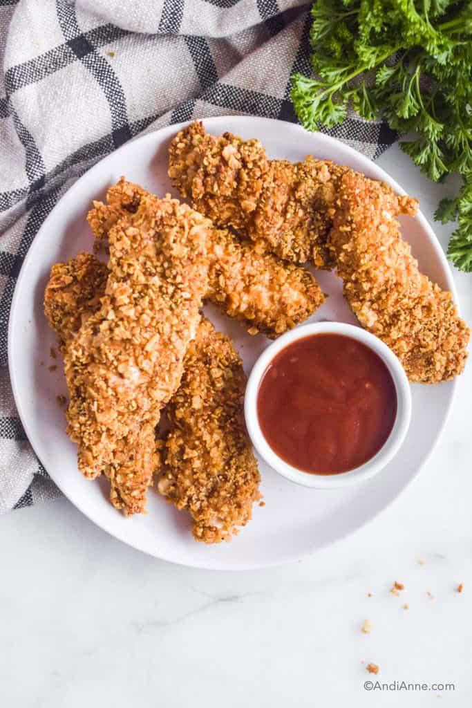 tortilla chip chicken tenders on a white plate with dipping sauce. parsley and kitchen towel beside it.