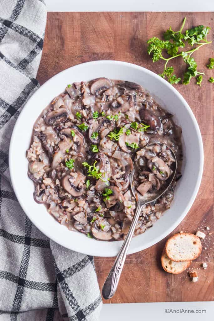 wild rice mushroom soup in a white bowl with large silver spoon. Parsley, croutons and a kitchen towel surround the white bowl.