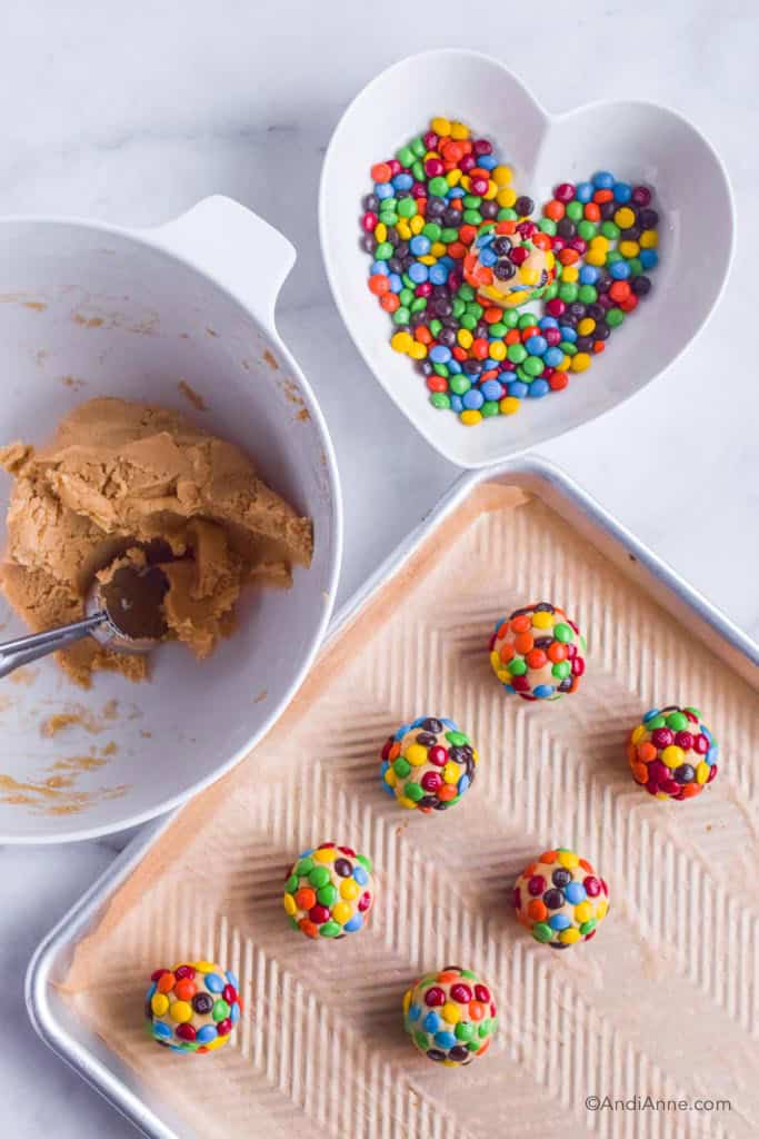 White bowl of batter with cookie scoop. Heart shaped bowl with mini M&Ms, cookie sheet with balls of cookie dough covered in M&Ms