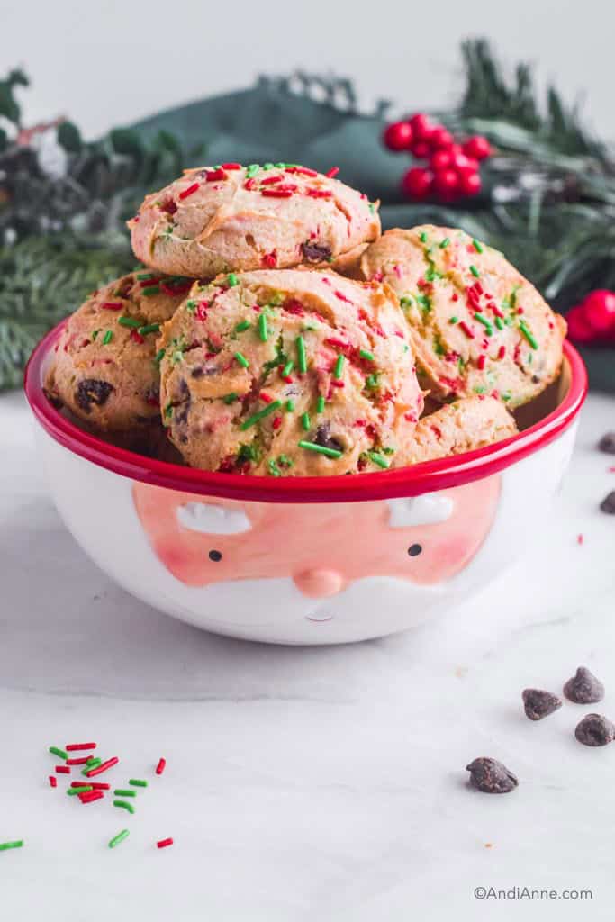 red and green sprinkled cake mix santa cookies piled in a santa designed bowl. Christmas greenery in the background and a few chocolate chips and sprinkles in the front.