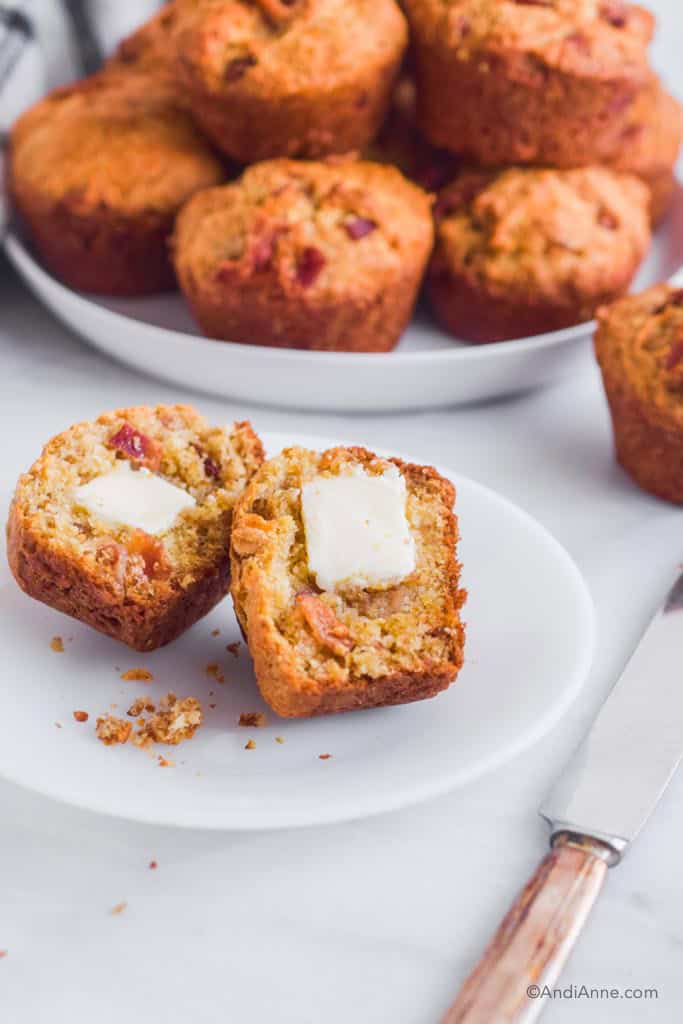One bacon cheese muffin biscuit sliced in half with butter. Plate of muffins in background.