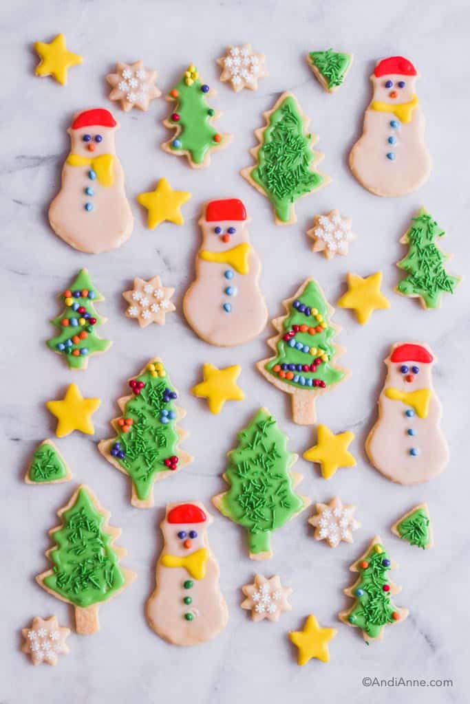 christmas sugar cookies on marble counter. Cookies include snowman shape, green trees with candy ornaments, mini snowflakes, and yellow stars.
