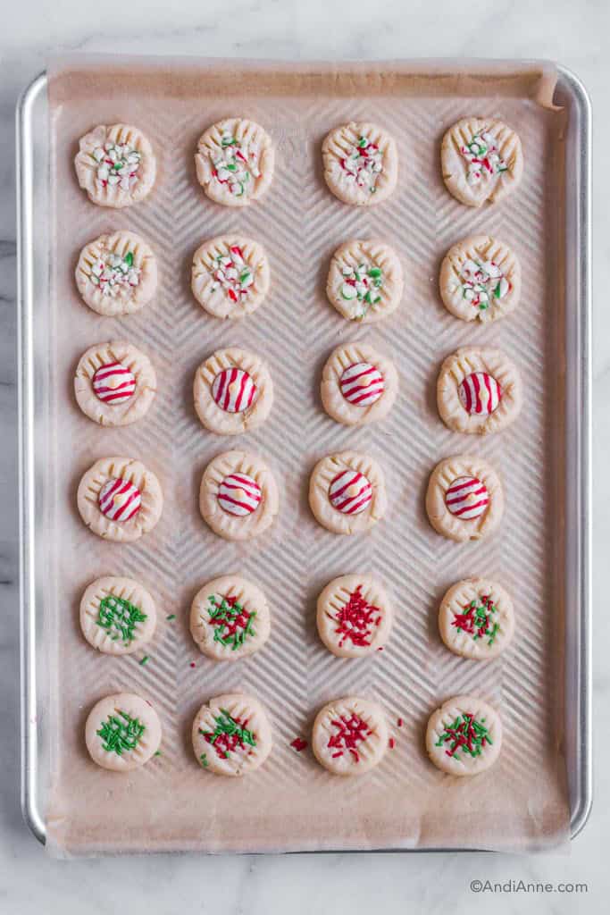 Baking sheet with parchment paper and whipped shortbread cookies with various christmas candies on top of each.