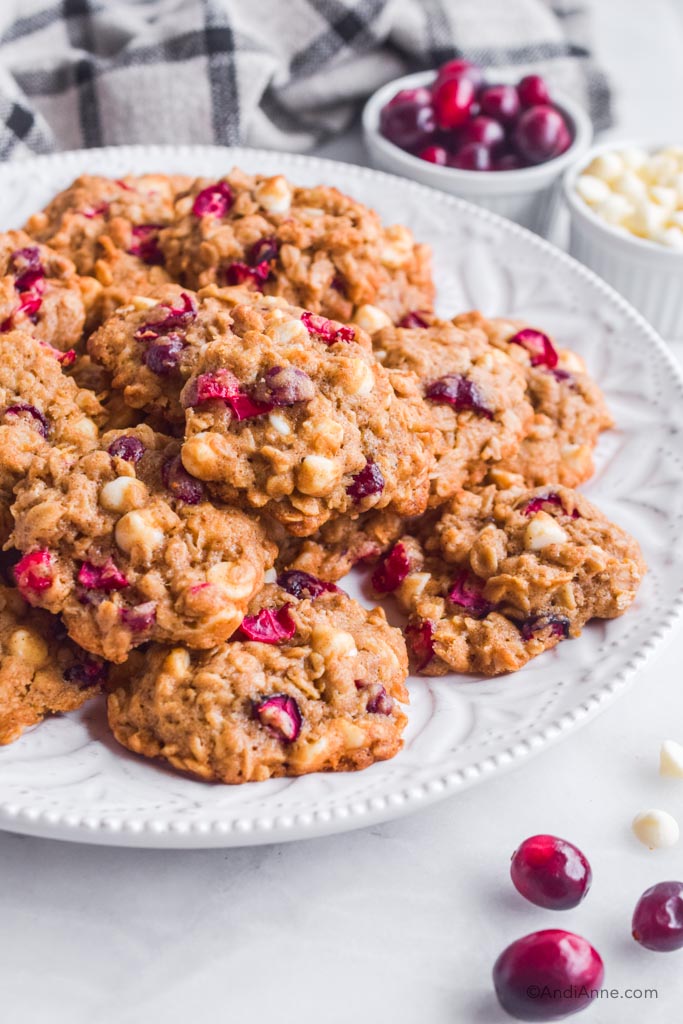 Oatmeal cranberry cookies with white chocolate chips stacked on a white plate with cranberries and chocolate chips in small bowls in the background.