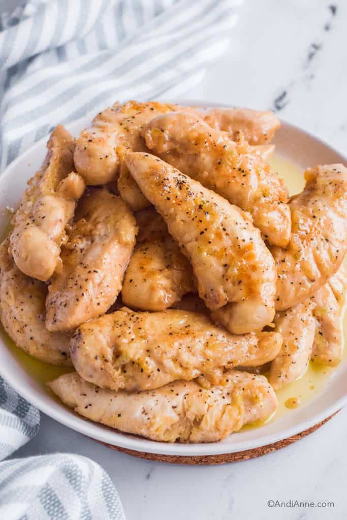pile of lemon garlic chicken tenders on white plate with striped kitchen towel behind it.