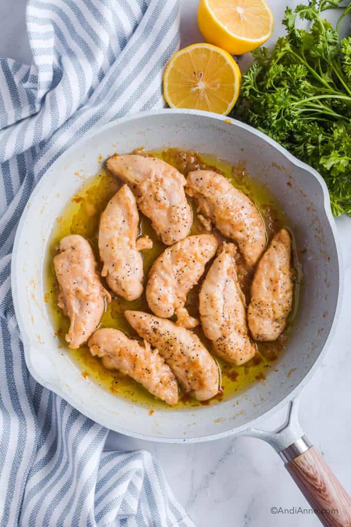 cooked maple lemon garlic chicken tenders in a frying pan with kitchen towel, lemon and parsley surrounding it.