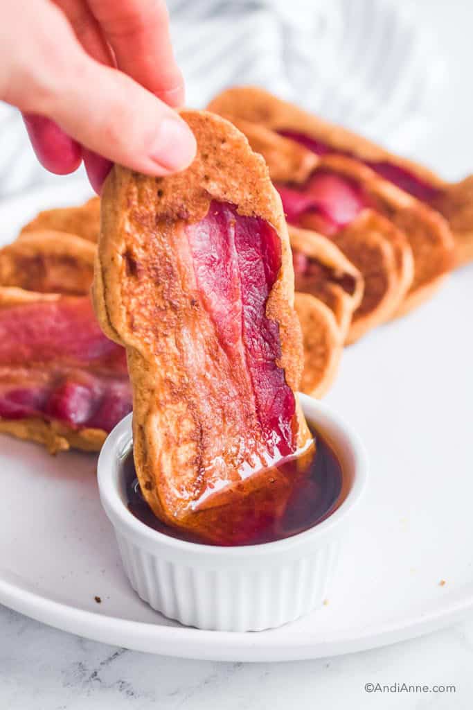 Hand dipping bacon pancakes in small bowl of maple syrup, with stack of pancakes behind it.