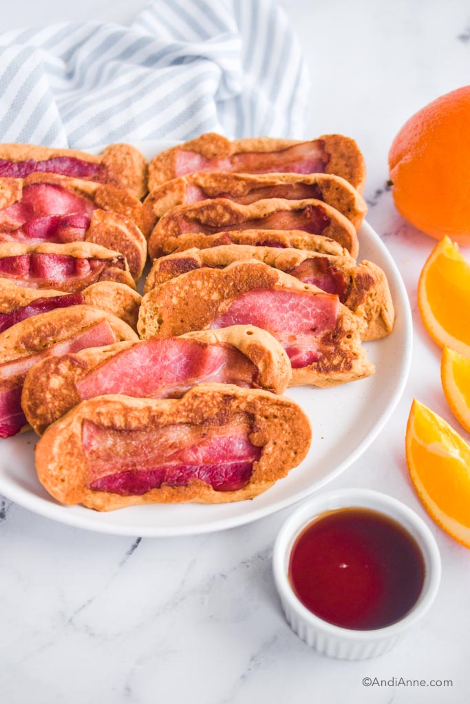 Bacon pancake dippers stacked on plate with maple syrup, orange slices and kitchen towel surrounding it.