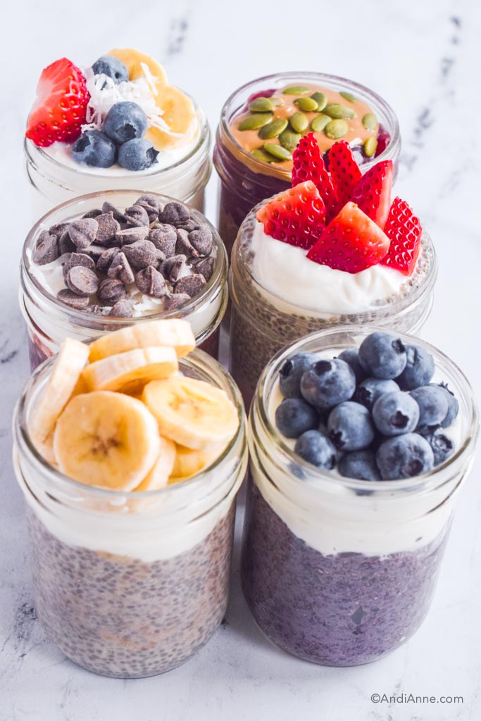 six chia pudding flavors in mason jars with various chopped fruit, chocolate chips and seeds on top.