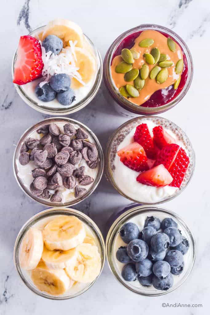 Looking down at tops of six chia puddings with chocolate chips, sliced strawberries, blueberries, bananas, nut butter, and pumpkin seeds.